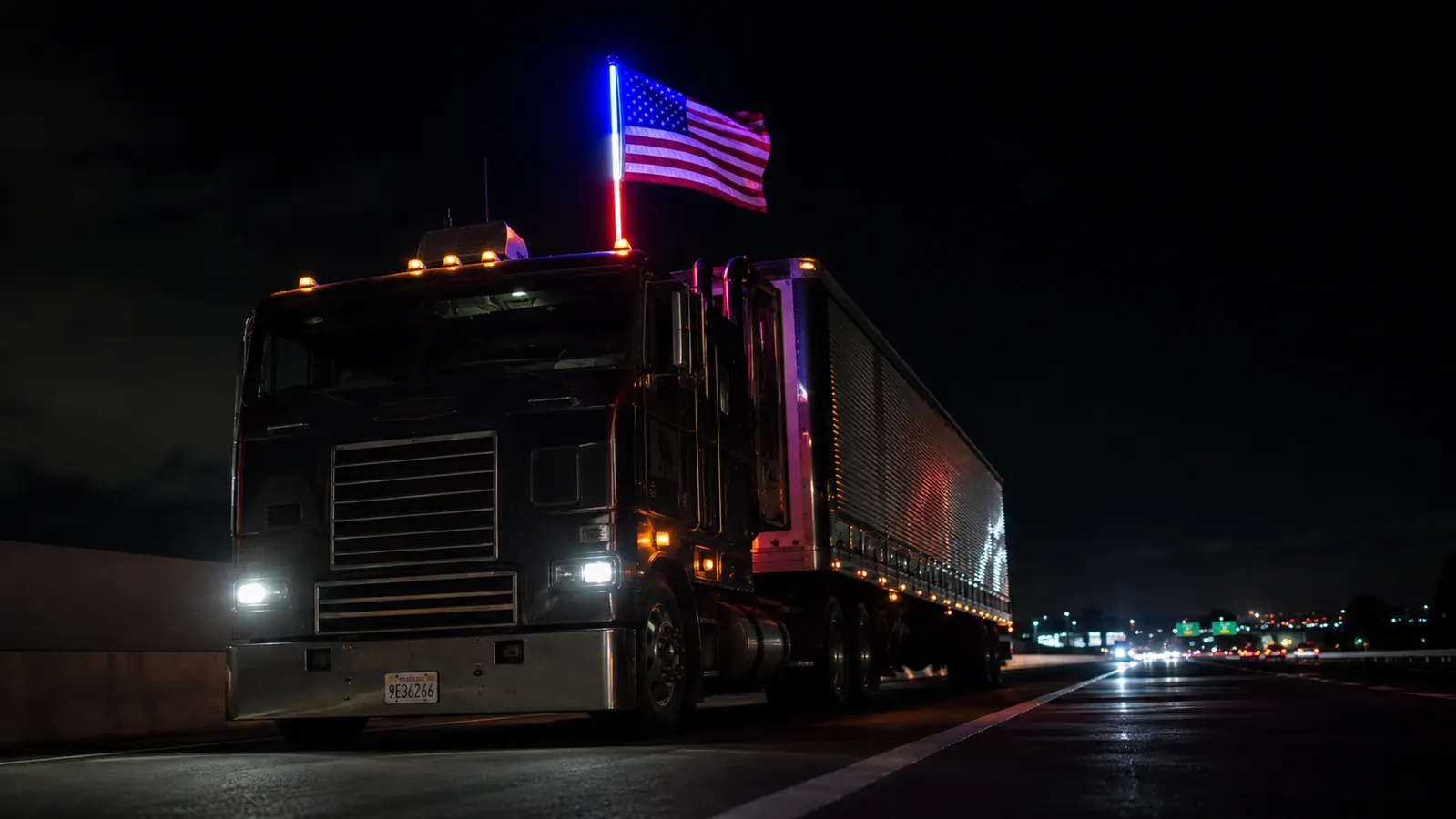 Cab-over semi truck on a dark interstate at night with an illuminated red, white, and blue LED whip flag pole glowing from the cab roof, highway stretching ahead